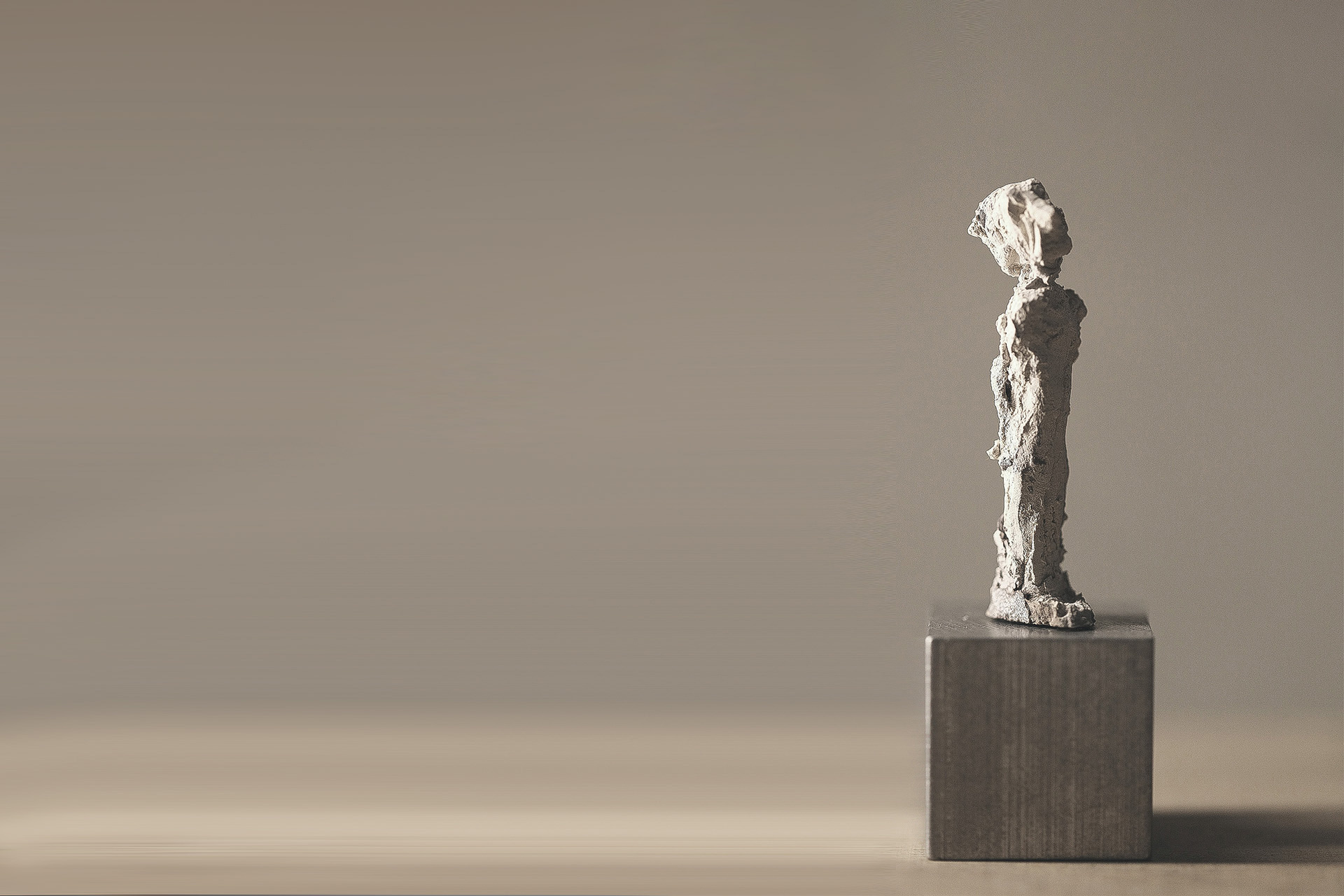 Clay sculpture of a narrow standing figure on a plinth in front
            of an empty light wall
