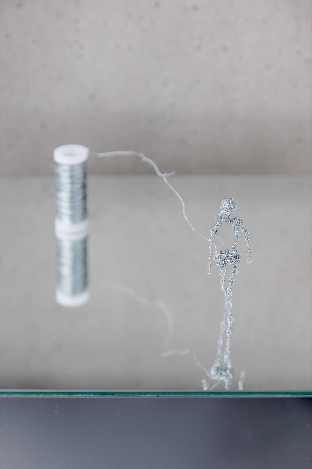 Wire sculpture on a glass plate: a wire runs off a reel
                  and becomes a standing figure